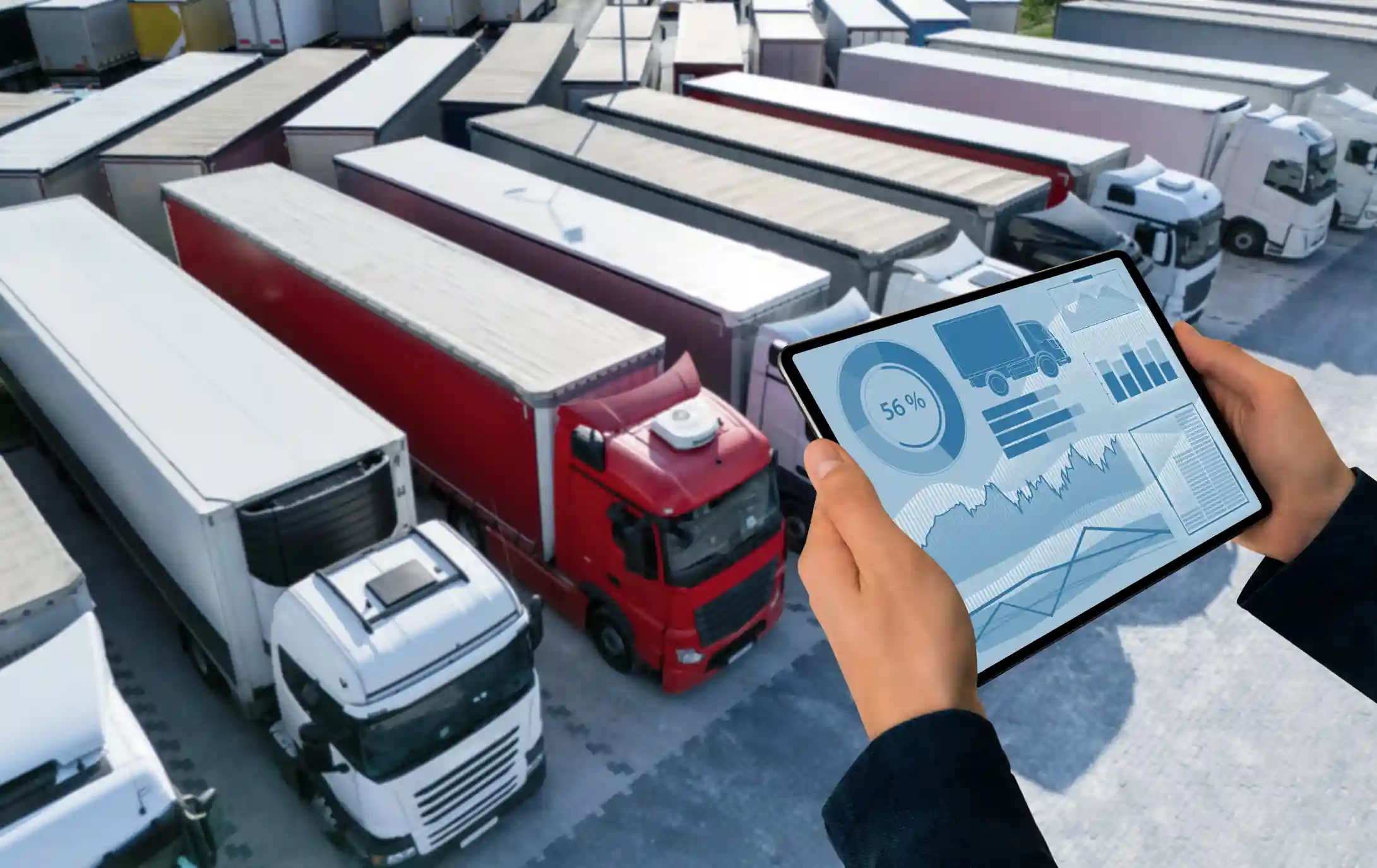 Manos de un gerente sosteniendo una tablet con dashboard de gestión de flota sobre el fondo de camiones estacionados